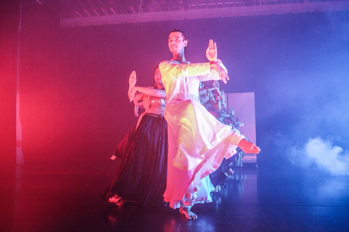 ABC company dancers in white and dark anarkalis moving in a line under red and blue stage light
